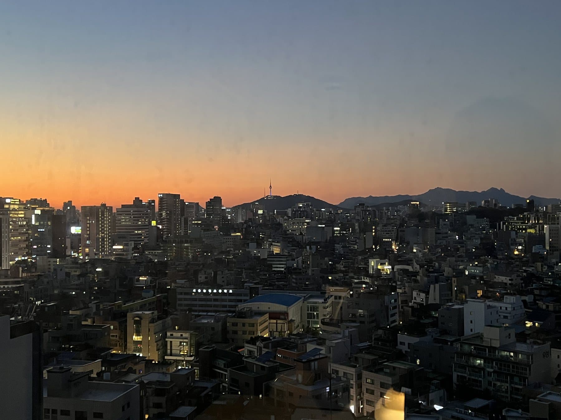 부트캠프룸 창밖으로 보이는 해질녘 서울 — 주황빛 하늘 아래 남산타워 실루엣과 그 뒤 북한산 능선